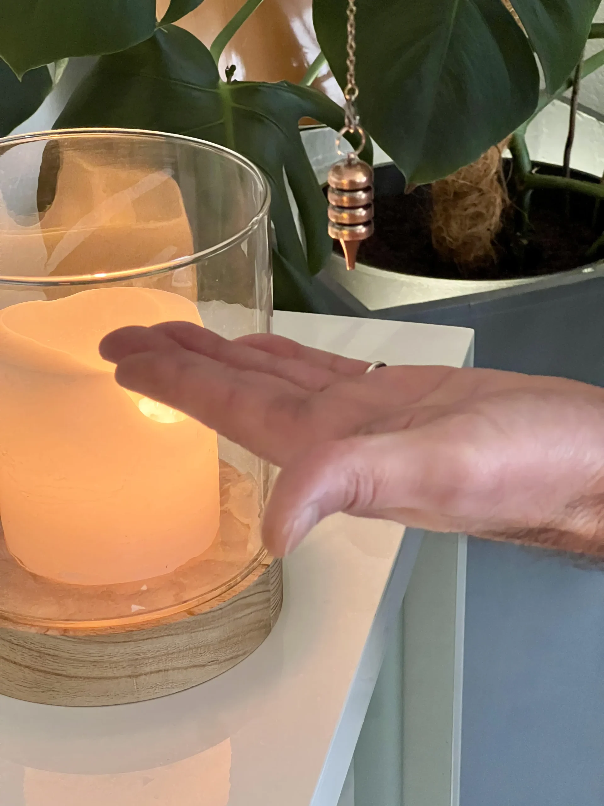soins radiesthesie holistique pendule bougie.webp scaled - Reiki près de Cholet & Nantes Pendule en cuivre en mouvement au-dessus d'une main, dans une ambiance lumineuse et apaisante.